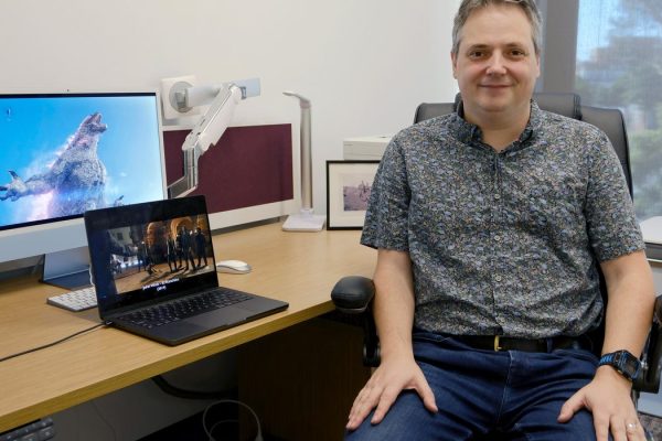 a white man in business casual attire seated at his desk with a laptop and desktop computer each playing a different movie on t