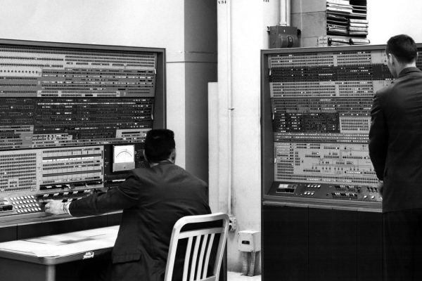 two men stand at 1960s computer consoles containing many buttons knobs dials and lights
