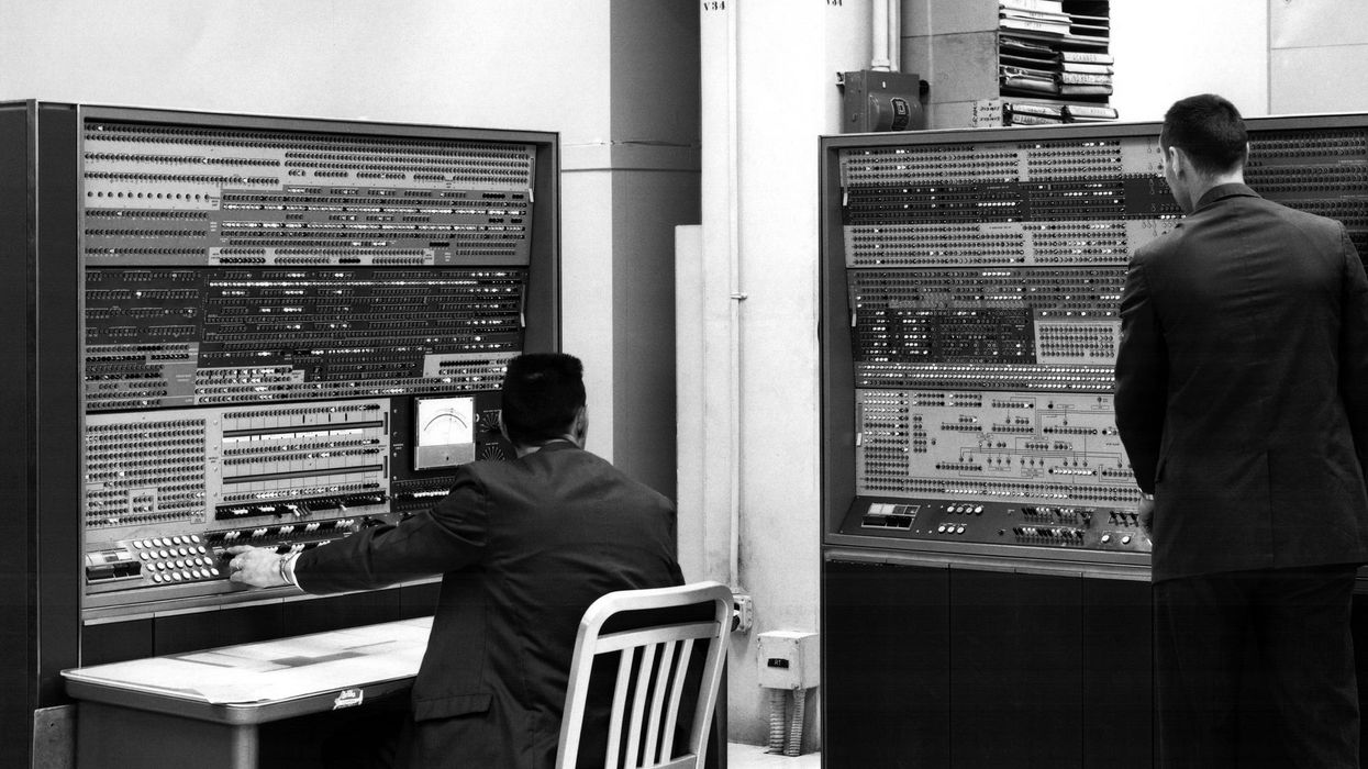 two men stand at 1960s computer consoles containing many buttons knobs dials and lights